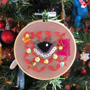 May include: A handmade Christmas ornament featuring an embroidered eye with sequins and beads in a circular wooden hoop.