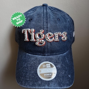 May include: A dark blue denim baseball cap with a white and red "Tigers" logo with rhinestones. The cap is adjustable and has a New Era logo on the side.