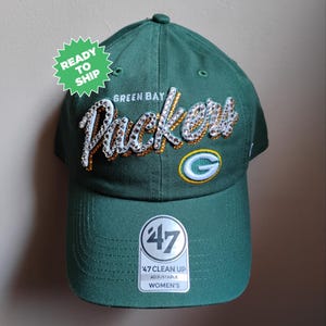 May include: Green adjustable baseball cap with the text "Green Bay Packers" in white and gold rhinestones. The cap has a curved brim and a small team logo on the side. The tag reads "47 Clean Up Adjustable Women's".