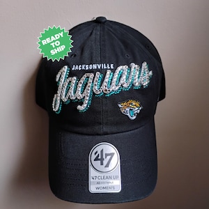 May include: Black adjustable baseball cap with the text "Jacksonville Jaguars" in silver and teal. The cap features the team logo and a "47 Clean Up" tag. A green sticker reads "Ready to Ship".