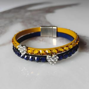 May include: Two stacked bracelets. The top bracelet is yellow with amber-colored rhinestones and a blue interior. The bottom bracelet is dark blue with blue rhinestones. Both bracelets have a silver heart charm.