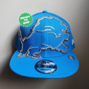 May include: A blue and silver baseball cap with a rhinestone Detroit Lions logo. The cap is a New Era 9Fifty Snapback.