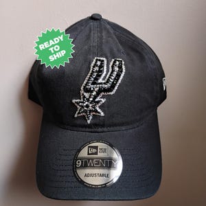 May include: Black adjustable 9TWENTY baseball cap with the San Antonio Spurs logo in silver and black rhinestones. The cap has a curved peak and a small New Era logo. A green sticker reads "READY TO SHIP".