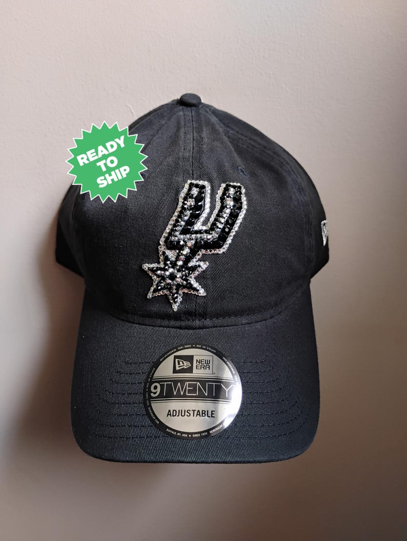 May include: Black adjustable 9TWENTY baseball cap with the San Antonio Spurs logo in silver and black rhinestones. The cap has a curved peak and a small New Era logo. A green sticker reads "READY TO SHIP".