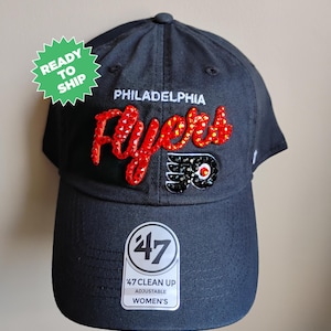 Può includere: Cappellino da baseball nero con un logo "Flyers" rosso e bianco con strass. Il logo include anche un logo Flyers nero e bianco con strass. Il cappello ha un'etichetta bianca "47 Clean Up Adjustable Women's".