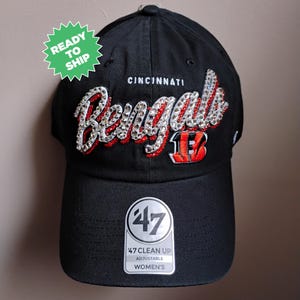 May include: Black adjustable baseball cap with the word "Bengals" in sparkling rhinestones and the word "Cincinnati" above it. The cap has a curved brim and a tag that says "47 Clean Up Adjustable Women's". A green sticker says "Ready to Ship".