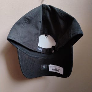 May include: A dark grey baseball cap with a curved peak and adjustable strap. The cap has a white tag and a small NBA logo sticker. The cap is hanging on a wall.