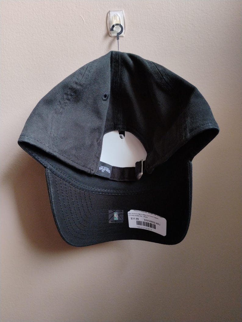 May include: A dark grey baseball cap with a curved peak and adjustable strap. The cap has a white tag and a small NBA logo sticker. The cap is hanging on a wall.