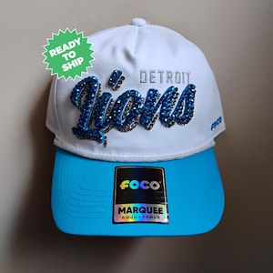 May include: White and blue Detroit Lions baseball cap with the team name in blue rhinestones. The brim is bright blue, and the cap has a FOCO tag. The text "DETROIT" is embroidered above the team name.