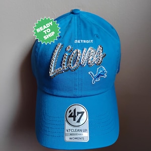 May include: A bright blue adjustable baseball cap with the text "Detroit Lions" in silver rhinestones. The cap features the team's logo and a "47 Clean Up" tag. The hat is ready to ship.