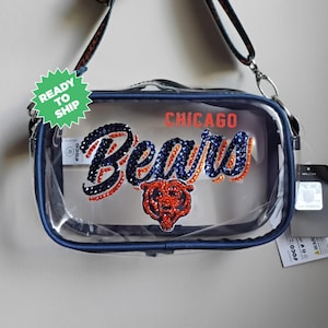 May include: Clear crossbody bag with navy blue trim and a navy blue and orange rhinestone design. The bag features the word "Bears" and the Chicago Bears logo. The bag has a detachable strap and a "Ready to Ship" sticker.