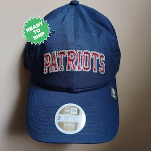 May include: A navy blue baseball cap with the word "PATRIOTS" in red and silver lettering. The cap has a curved brim and a small logo on the side. The tag on the cap says "9TWENTY WOMEN ADJUSTABLE".