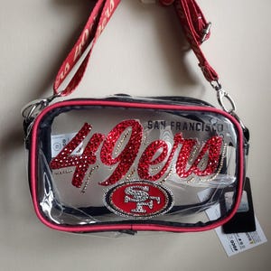 May include: Clear crossbody bag with red trim and a red and silver "49ers" logo. The bag has a red strap with white text and a team logo patch. The words "San Francisco" are above the logo.