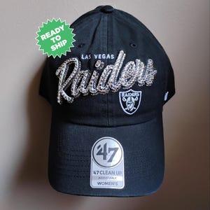 May include: Black adjustable baseball cap featuring the Las Vegas Raiders logo in silver rhinestones. The cap includes the Raiders shield logo and the text "47 Clean Up Adjustable Women's". A green "Ready to Ship" sticker is attached.