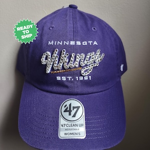 May include: A purple baseball cap featuring the text Minnesota Vikings Est. 1961. The word Vikings is embellished with clear rhinestones and a gold rhinestone line underneath. A 47 brand sticker is on the brim, and a green Ready To Ship tag is visible.