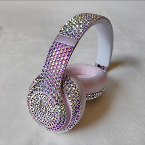 May include: Light pink over-ear headphones featuring a dense, sparkling layer of iridescent rhinestones covering the outer headband and ear cups. The Beats logo is visible on the side of the ear cup, surrounded by the reflective crystal embellishments.