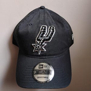 May include: Black adjustable baseball cap with the San Antonio Spurs logo in silver and black sequins. The cap has a curved peak and a 9TWENTY adjustable sticker. The New Era logo is visible on the side.
