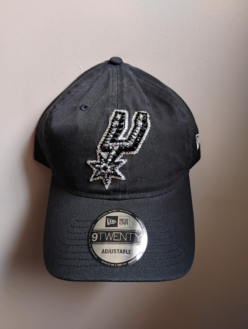 May include: Black adjustable baseball cap with the San Antonio Spurs logo in silver and black sequins. The cap has a curved peak and a 9TWENTY adjustable sticker. The New Era logo is visible on the side.