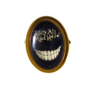 Peut inclure: Une épingle ovale dorée avec un fond noir et une bouche souriante blanche. Le texte "We're All Mad Here" est écrit en blanc sur le fond noir.