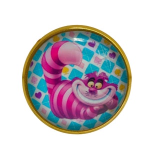 Peut inclure: Un cabochon rond en verre à bordure dorée présente une image de dessin animé du Chat du Cheshire d'Alice au pays des merveilles. Le chat est rose et violet avec un large sourire et est placé sur un fond bleu et blanc avec un motif en losange.