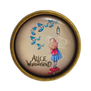 Peut inclure: Une épingle ronde en métal doré avec un dôme en verre représentant une illustration colorée d'Alice d'Alice au pays des merveilles tenant une bouteille et entourée de papillons bleus. Le texte "Alice au pays des merveilles" est imprimé sous l'image.