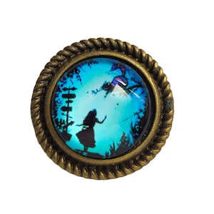 Peut inclure: Une bague en bronze avec un camée en verre bleu représentant une silhouette d'Alice d'Alice au pays des merveilles tombant dans les airs. Le camée est entouré d'un motif en forme de corde.