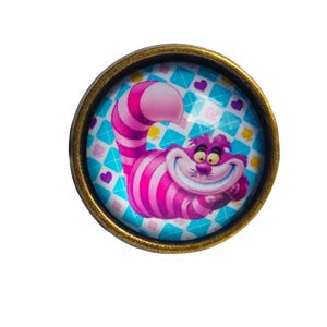 May include: A round, gold-colored pin with a glass dome featuring a cartoon image of the Cheshire Cat from Alice in Wonderland. The cat is pink and purple with a wide grin and is set against a blue and white background with a diamond pattern.