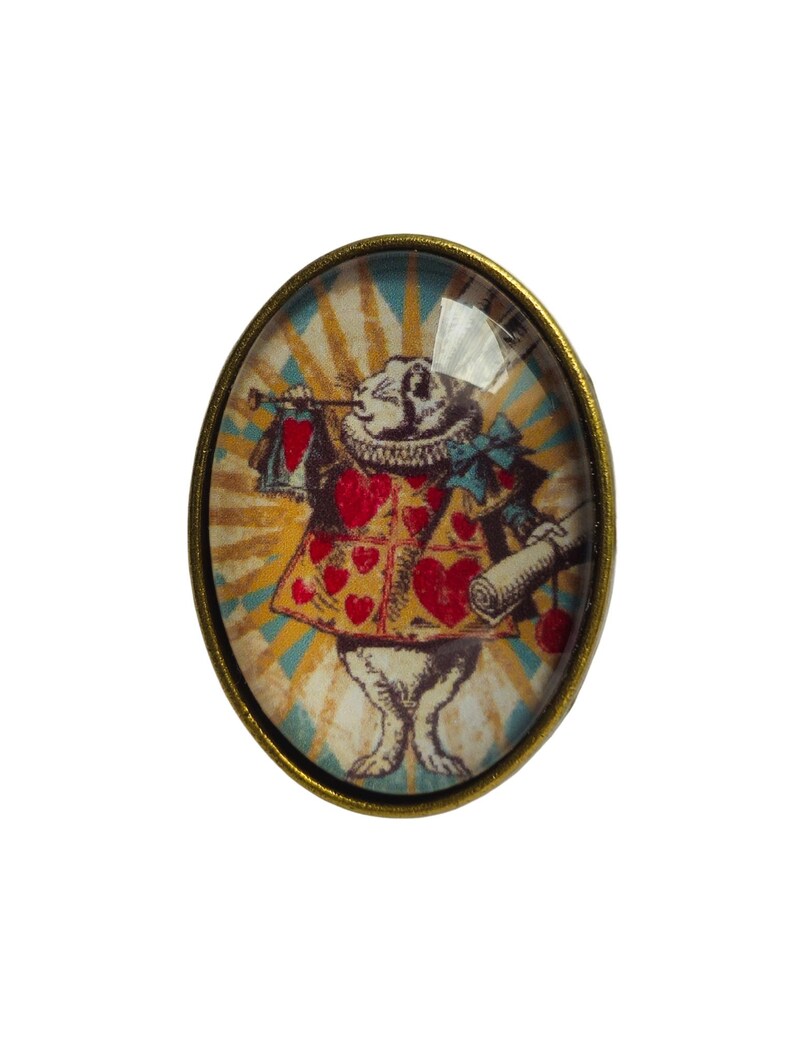 May include: Oval-shaped glass cabochon with a gold-toned metal frame. The cabochon features a vintage illustration of the White Rabbit from Alice in Wonderland, wearing a red heart-shaped waistcoat and holding a scroll and a trumpet.