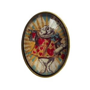 Peut inclure: Cabochon en verre de forme ovale avec un cadre en métal doré. Le cabochon présente une illustration vintage du Lapin Blanc d'Alice au Pays des Merveilles, portant un gilet rouge en forme de cœur et tenant un rouleau et une trompette.