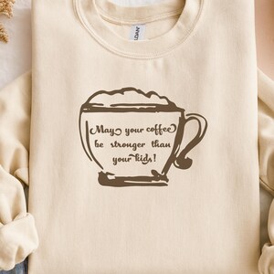 May include: A cream-colored sweatshirt with the phrase "May your coffee be stronger than your kids!" printed in brown within a coffee cup graphic. The long-sleeved top is folded, showing the cuffs and neckline.