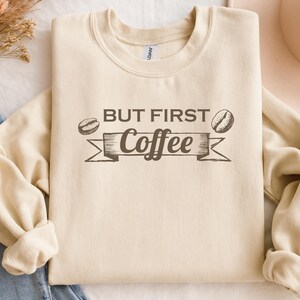 May include: Cream-colored sweatshirt with the phrase "BUT FIRST Coffee" in dark brown lettering. The design includes coffee bean illustrations. The sweatshirt is folded, with the sleeves rolled up, and is placed on a white surface.