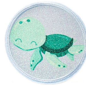 Könnte beinhalten: Ein weißer, glitzernder, runder Patch mit einer grünen und blauen Comic-Schildkröte mit einem lächelnden Gesicht.