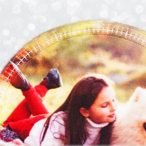 May include: A circular patch showing a girl with long brown hair and a white turtleneck, lying next to a fluffy white dog. The girl wears red trousers and black shoes. The patch has a stitched border and a blurred background.