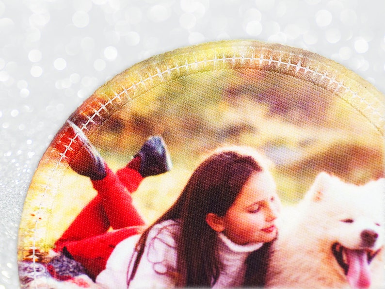 May include: A circular patch showing a girl with long brown hair and a white turtleneck, lying next to a fluffy white dog. The girl wears red trousers and black shoes. The patch has a stitched border and a blurred background.