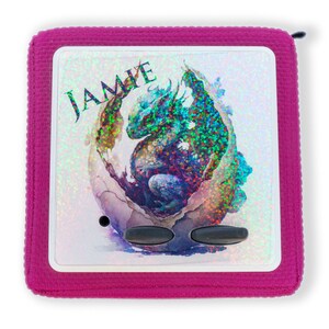 Könnte beinhalten: Eine rosa Stofftasche mit einem durchsichtigen Plastikfenster. Das Fenster zeigt ein holografisches Bild eines blauen Drachen, der aus einem Ei schlüpft. Der Name "JAMIE" ist in schwarzen Buchstaben über dem Drachen gedruckt.