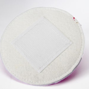 May include: A circular, white, felt-like pad with a square, white, velcro patch in the centre. The pad has a stitched edge and a light purple border. The background is white.