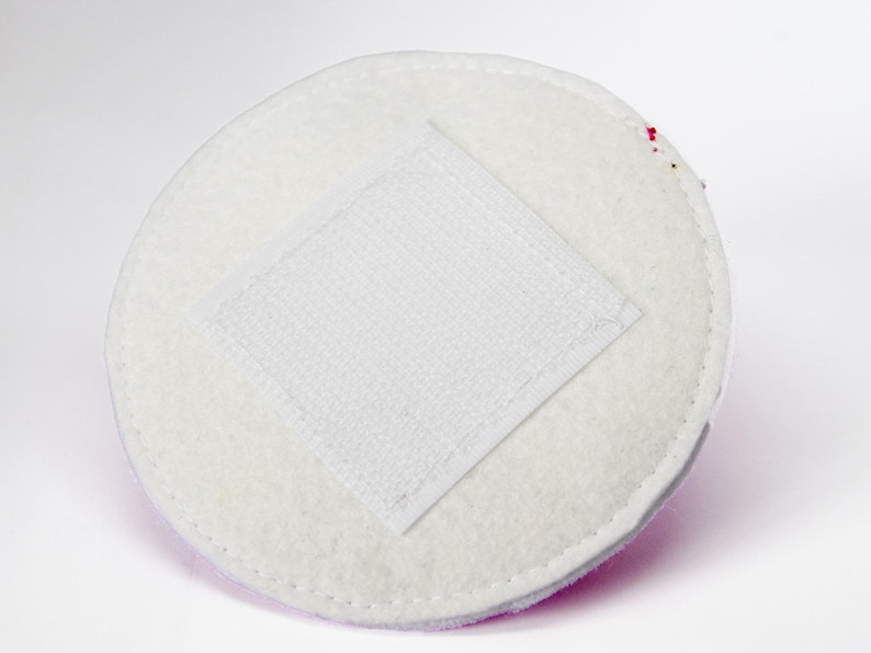 May include: A circular, white, felt-like pad with a square, white, velcro patch in the centre. The pad has a stitched edge and a light purple border. The background is white.