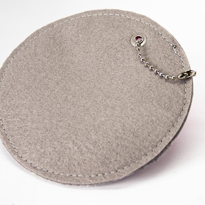 May include: A grey felt coaster with a white stitched border and a silver ball chain attached to a metal grommet. The coaster is round with a slightly irregular shape.