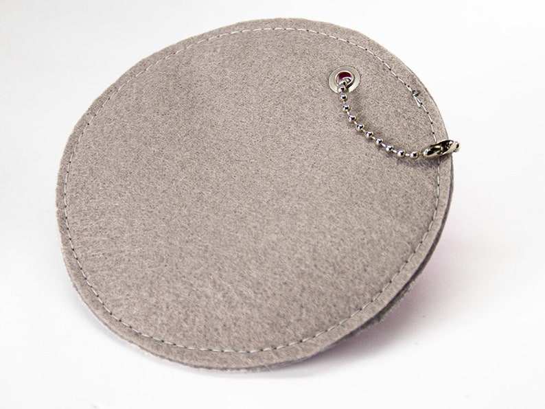 May include: A grey felt coaster with a white stitched border and a silver ball chain attached to a metal grommet. The coaster is round with a slightly irregular shape.