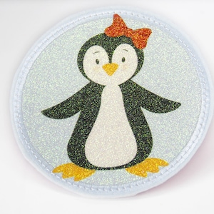 Könnte beinhalten: Ein weißer, runder, glitzernder Patch mit einem schwarz-weißen Pinguin, der eine rote Schleife trägt. Der Pinguin hat einen gelben Schnabel und orangefarbene Füße.