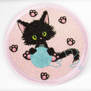 Könnte beinhalten: Ein schwarzes Kätzchen mit grünen Augen spielt mit einem blauen Wollknäuel auf einem rosa Glitzerhintergrund. Das Kätzchen ist von rosa Pfotenabdrücken umgeben.