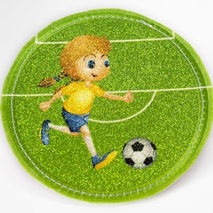 Könnte beinhalten: Ein grüner Glitzer-Stoff-Patch mit einem weißen, gestickten Rand. Der Patch zeigt ein Cartoon-Mädchen, das auf einem grünen Feld mit weißen Linien einen Fußball tritt.
