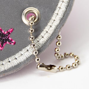 May include: Close-up of a grey, star-shaped accessory with pink and black embroidered stars. It features a silver chain and a silver metal grommet. The edge is trimmed with silver stitching.