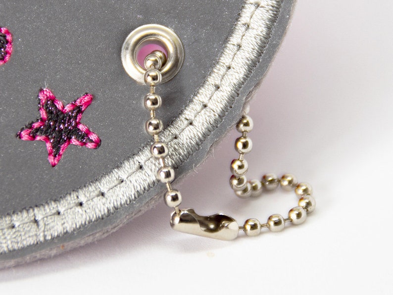 May include: Close-up of a grey, star-shaped accessory with pink and black embroidered stars. It features a silver chain and a silver metal grommet. The edge is trimmed with silver stitching.