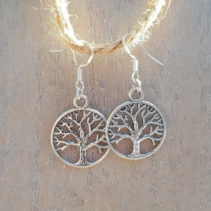 Op de afbeelding: Een paar zilverkleurige oorbellen met een levensboommotief. Elke oorbel heeft een cirkelvormige hanger met een gedetailleerd boomontwerp. De oorbellen hangen aan zilverkleurige oorhaken en worden getoond tegen een houten achtergrond.