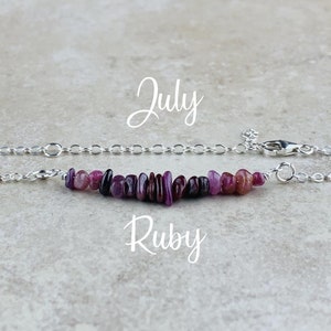 May include: A silver chain necklace with a bar of ruby gemstones. The word "July" is written above the necklace, and the word "Ruby" is written below it. The gemstones are various shades of red and purple.