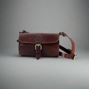 Op de afbeelding: Bruine leren crossbodytas met een rechthoekige vorm. De tas heeft een klepsluiting met een goudkleurige gesp en een lange, verstelbare riem. Het leer heeft een getextureerde afwerking.