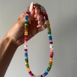 Puede incluir: Un collar de cuentas colorido que presenta un espectro de tonos, incluyendo rosa, rojo, naranja, amarillo, verde, azul y morado. El collar tiene una cadena y un cierre de color dorado. Las cuentas son de tamaño y forma uniformes.
