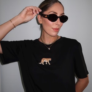 May include: Black t-shirt with a leopard graphic. The shirt is paired with leopard print pants and brown oval sunglasses. A gold necklace and bracelet complete the look. The sunglasses have a tortoiseshell pattern.