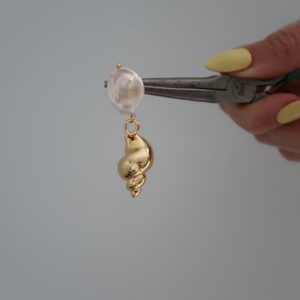 May include: A gold seashell charm with a white pearl attached to it. The charm is being held by a pair of pliers.